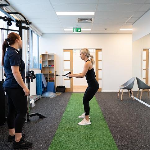 [In A Gym Setting, Clinican Is Talking To The Patient And The Patient Is Using The Cable Machine In A Row Position And Half Squatting, The Patient Is Wearing All Black And Pink Shoes, The Clinician Is Standing To The Left Of The Patient, Wearing A Navy Polo Sports & Spinal Shirt With Black Pants And Black Shoes]